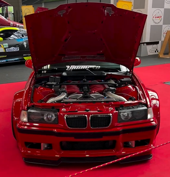 Unlimited Auto's 1000 PS BMW E36 BiTurbo V12 build with widebody kit and open hood at an event.